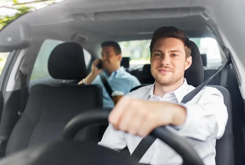 A driver and a rear-seat passenger travelling in a car, both wearing fastened seatbelts.