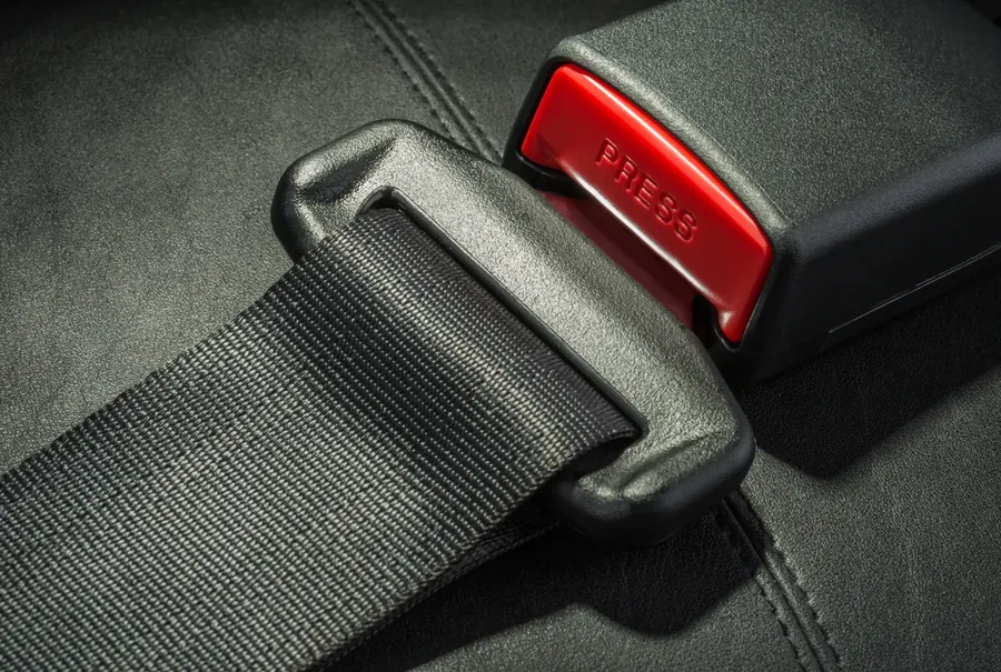 Seatbelt webbing feeding through a moulded latch plate next to a red-release buckle housing on a dark leather seat.