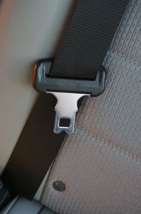 A black seatbelt with its metal latch plate resting against a light grey fabric seat back.